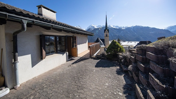A building with a sloped roof, a wooden door, and a window. There is a paved pathway leading to the entrance.