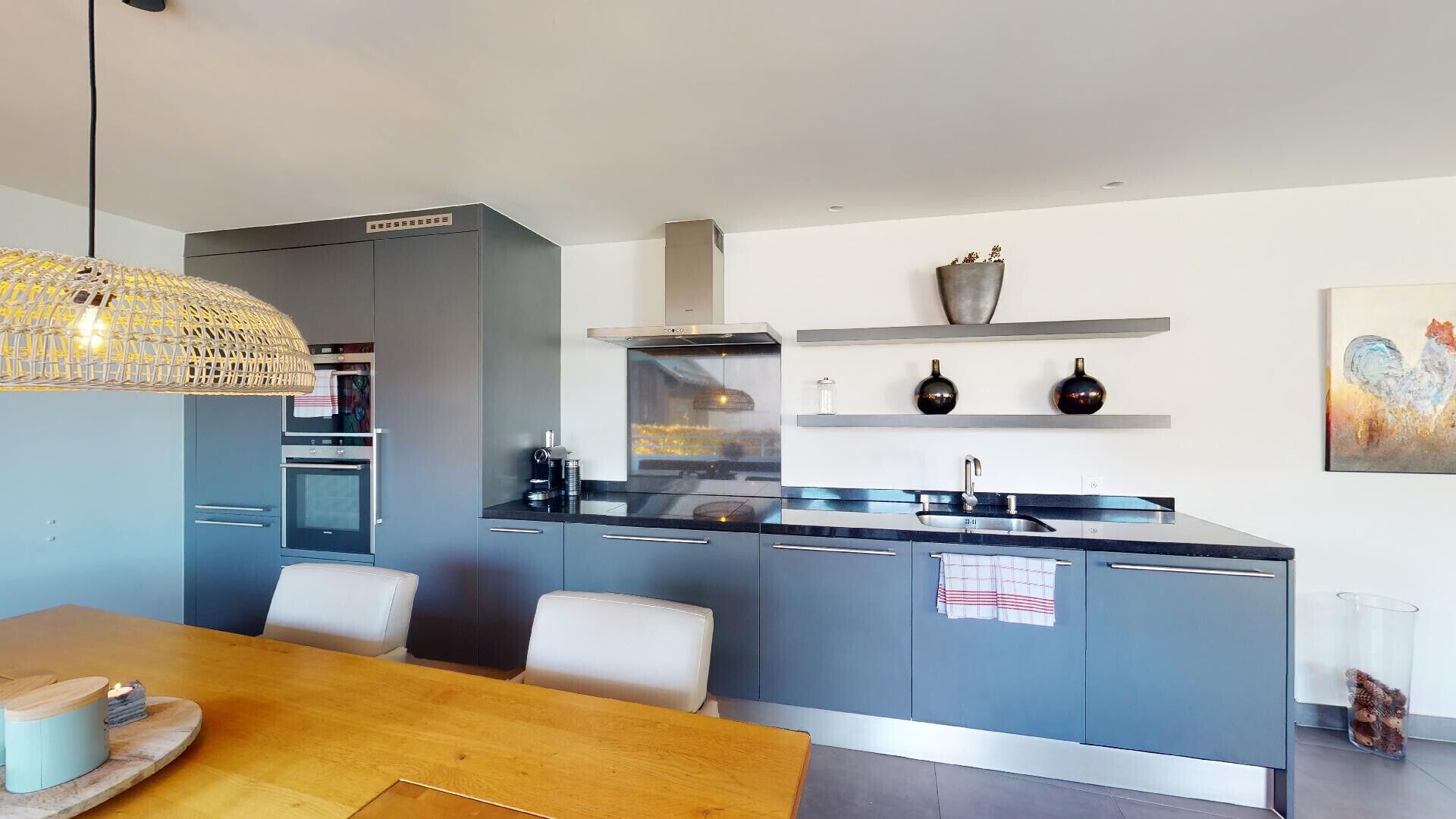 A modern kitchen with grey cabinets, a stainless steel range hood, a built-in oven, and a dining area with a wooden table and chairs.
