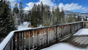 Una terraza de madera cubierta de nieve, rodeada de árboles nevados y un paisaje montañoso.