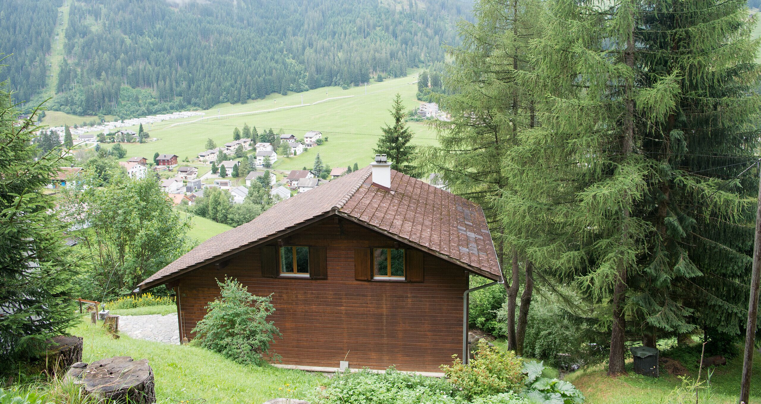 A wooden cabin with a sloped roof surrounded by dense forest and a distant village.
