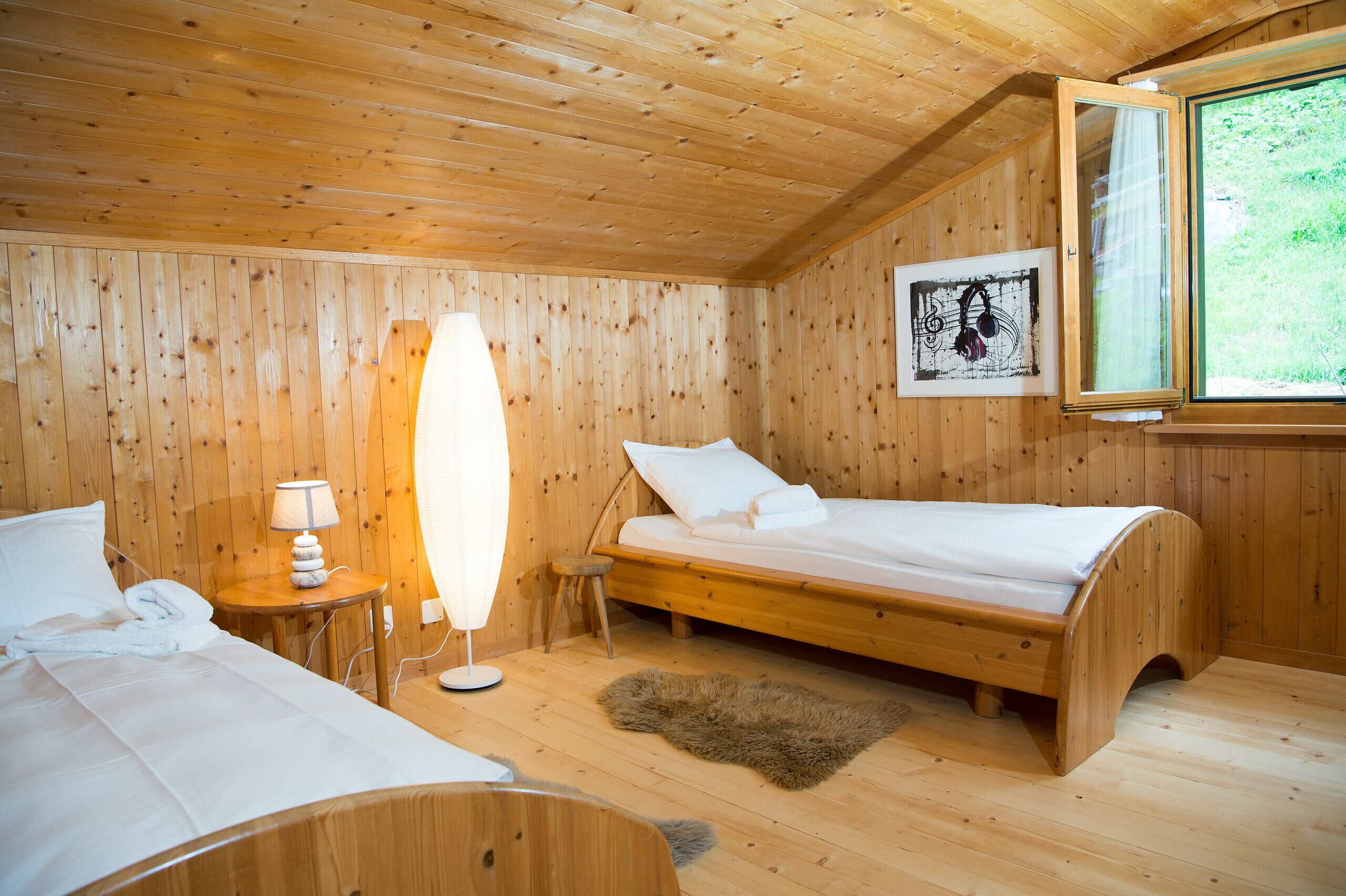 Un dormitorio de cabaña de madera con una cama, una mesita de noche con lámpara, un taburete pequeño y una alfombra de piel en el suelo.