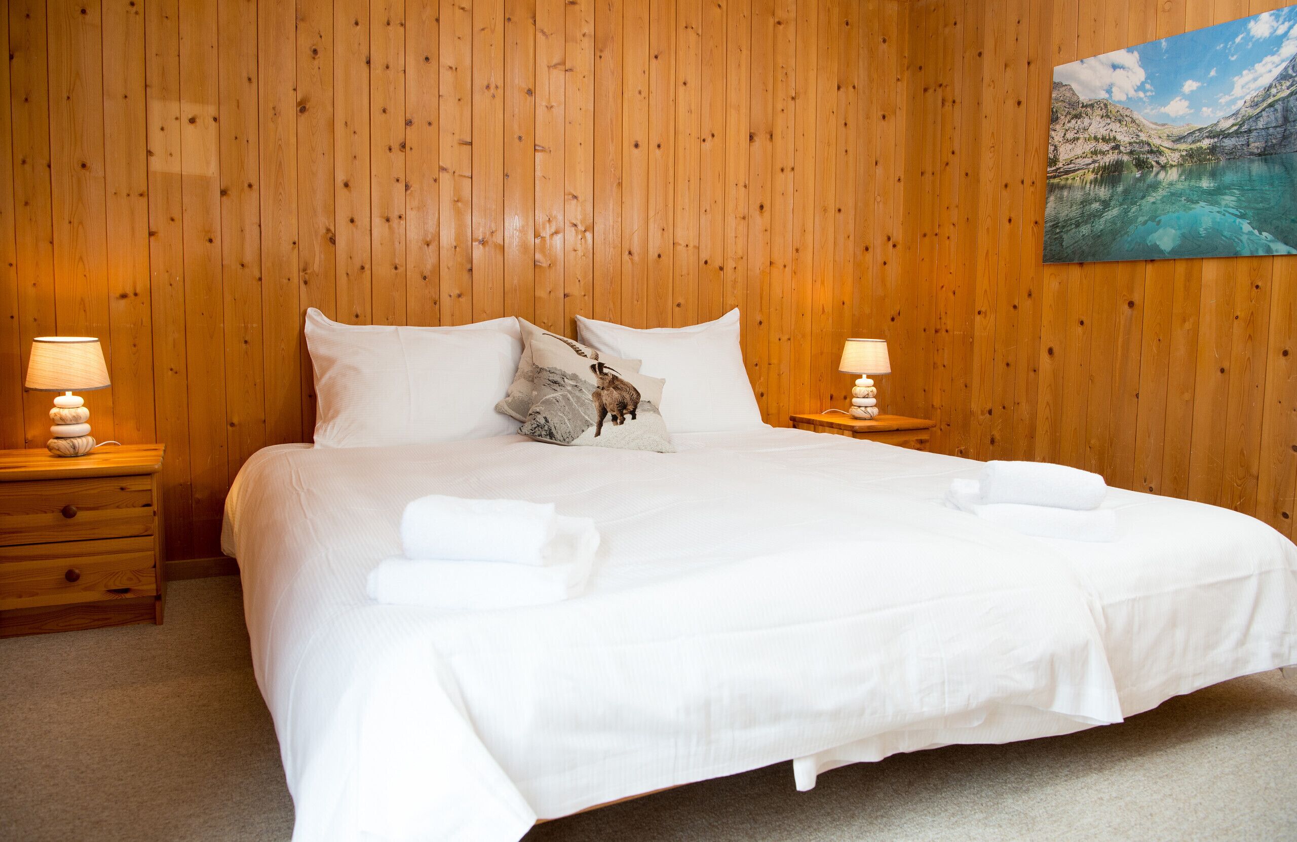 Una cama bien hecha con sábanas y almohadas blancas, dos mesitas de noche con lámparas, y un cuadro de un paisaje montañoso en la pared.