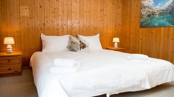Una cama bien hecha con sábanas y almohadas blancas, dos mesitas de noche con lámparas, y un cuadro de un paisaje montañoso en la pared.