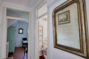 Ein Flur mit einer weißen Tür, die zu einem Zimmer mit einem Bett und einem Sessel führt. An der Wand hängt ein gerahmtes Bild.