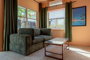 Una habitación con sofá, una mesita, ventana con cortinas y un cuadro en la pared.