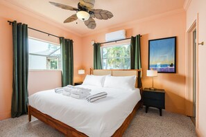 Habitación de hotel con cama, ventilador de techo, ventana con cortinas, un cuadro en la pared y una mesita de noche con lámpara.