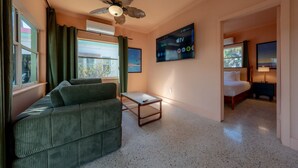 A hotel room with a sofa, a small table, a bed, a ceiling fan, a TV, and a window with curtains.