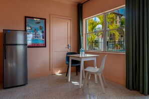 Una kitchenette con refrigerador, una mesa pequeña con sillas, una puerta y una ventana con vista a palmeras y edificios.