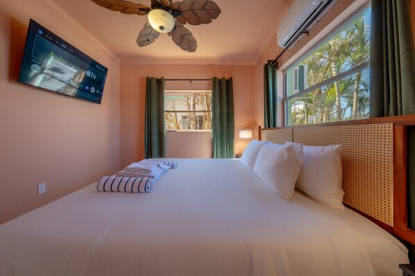 Una habitación de hotel con cama, ventilador de techo, televisor montado en la pared y ventana con cortinas.