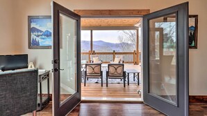 A room with a wooden floor, a television, and a door leading to an outdoor deck with chairs and a table. - Mountainside Family Retreat w/ Deck in Banner Elk! (Banner Elk)