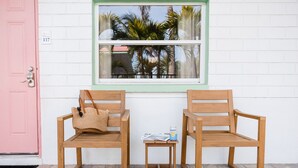 Two wooden chairs, a small table, a pink door, and a window with a view of palm trees.