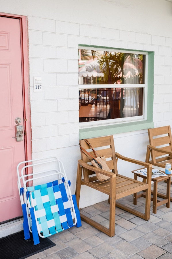A pink door with a keypad lock, a window with a view of palm trees, and two wooden chairs with a small table.