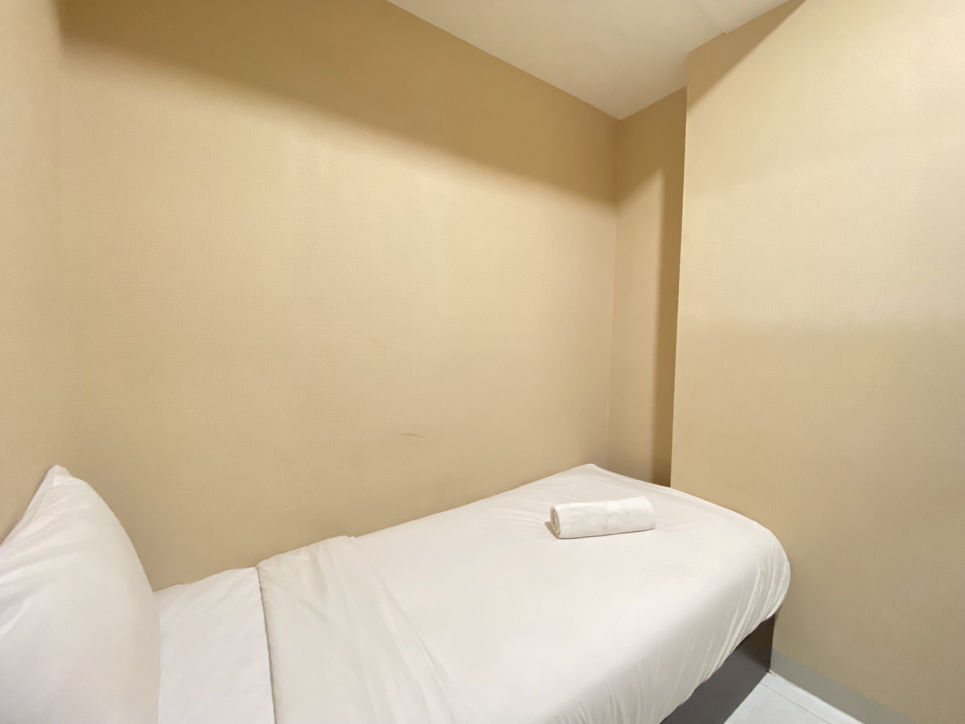 A small, single-bed room with a plain beige wall and a white bedspread.