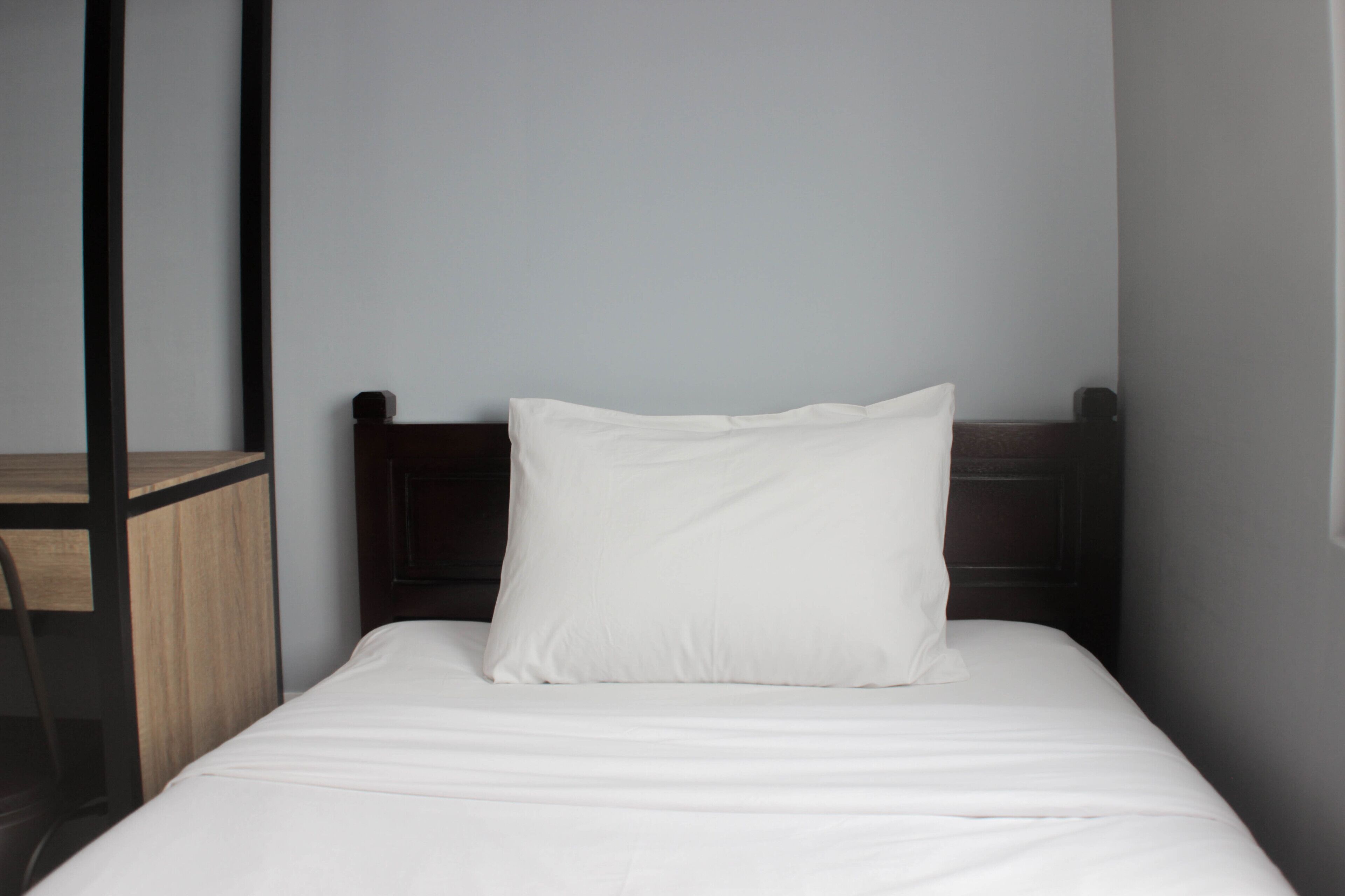A single bed with a white pillow and a wooden headboard in a room with a plain wall and a small wooden table to the left.