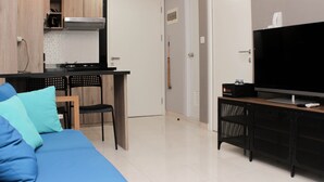 A modern living room with a blue sofa, a TV on a stand, a kitchen area with cabinets and a stove, and a door with a nameplate.