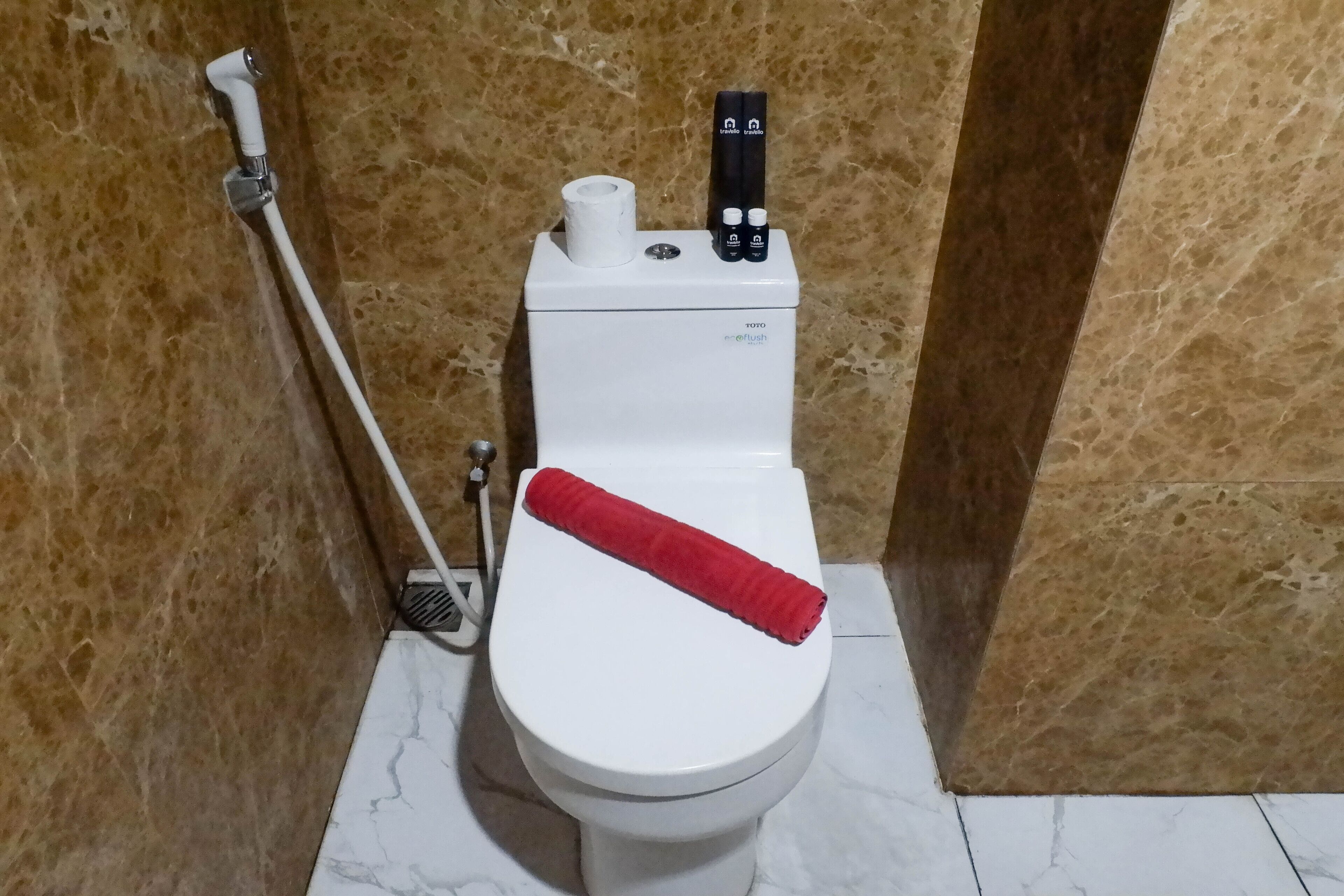 A toilet with a red towel, a toilet paper holder, and a bidet attachment on a marble floor.