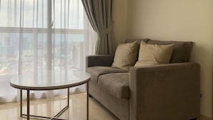 A modern living room with a sofa and a small round table, large windows with curtains, and a city view.