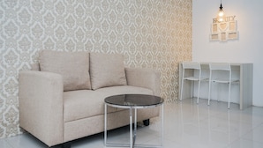 A beige sofa, a round glass table, and two white chairs in a room with patterned wallpaper and a hanging light fixture.