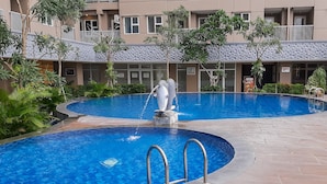 A swimming pool with a dolphin statue, surrounded by greenery and a building with balconies.