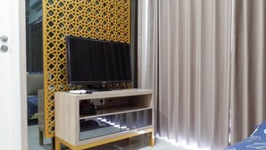 A room with a TV on a wooden stand, a glass cabinet, and a bed with a blue bedspread. - Comfy And Modern Wonderful 2Br Apartment At Tanglin Supermall Mansion (Surabaya)