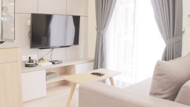 A modern living room with a flat-screen TV mounted on the wall, a wooden coffee table, a beige sofa, and a window with grey curtains. - Serene And Cozy 2Br At Meikarta Apartment (Cikarang)
