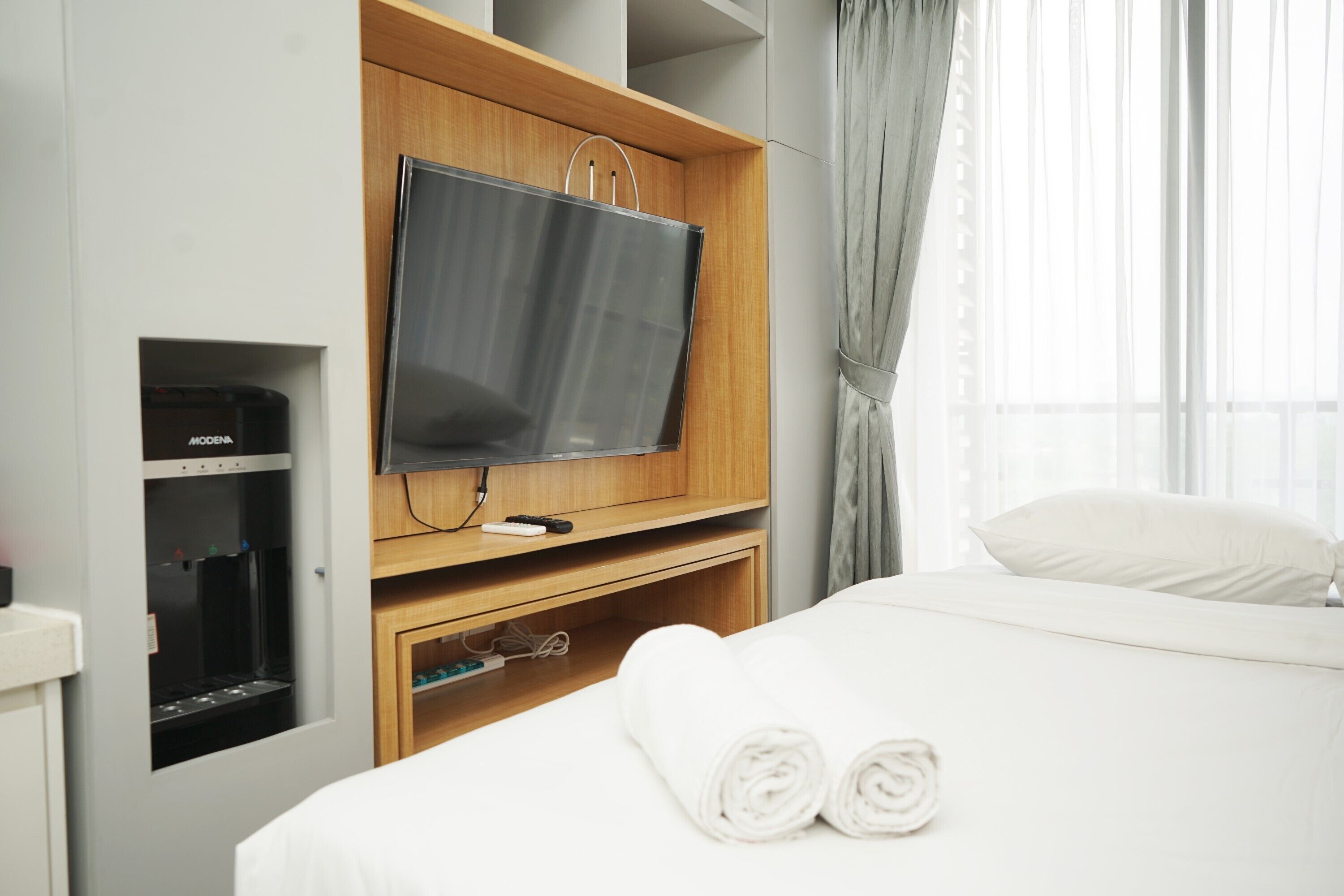 Habitación de hotel con cama, televisor montado en un mueble de madera, minibar y ventana con cortinas.
