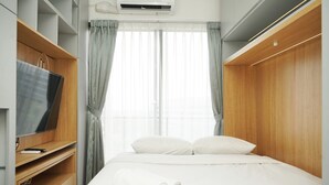 A hotel room with a bed, a TV on a wooden stand, and a window with curtains. - Minimalist And Cozy Studio At Sky House Bsd Apartment (Pagedangan)