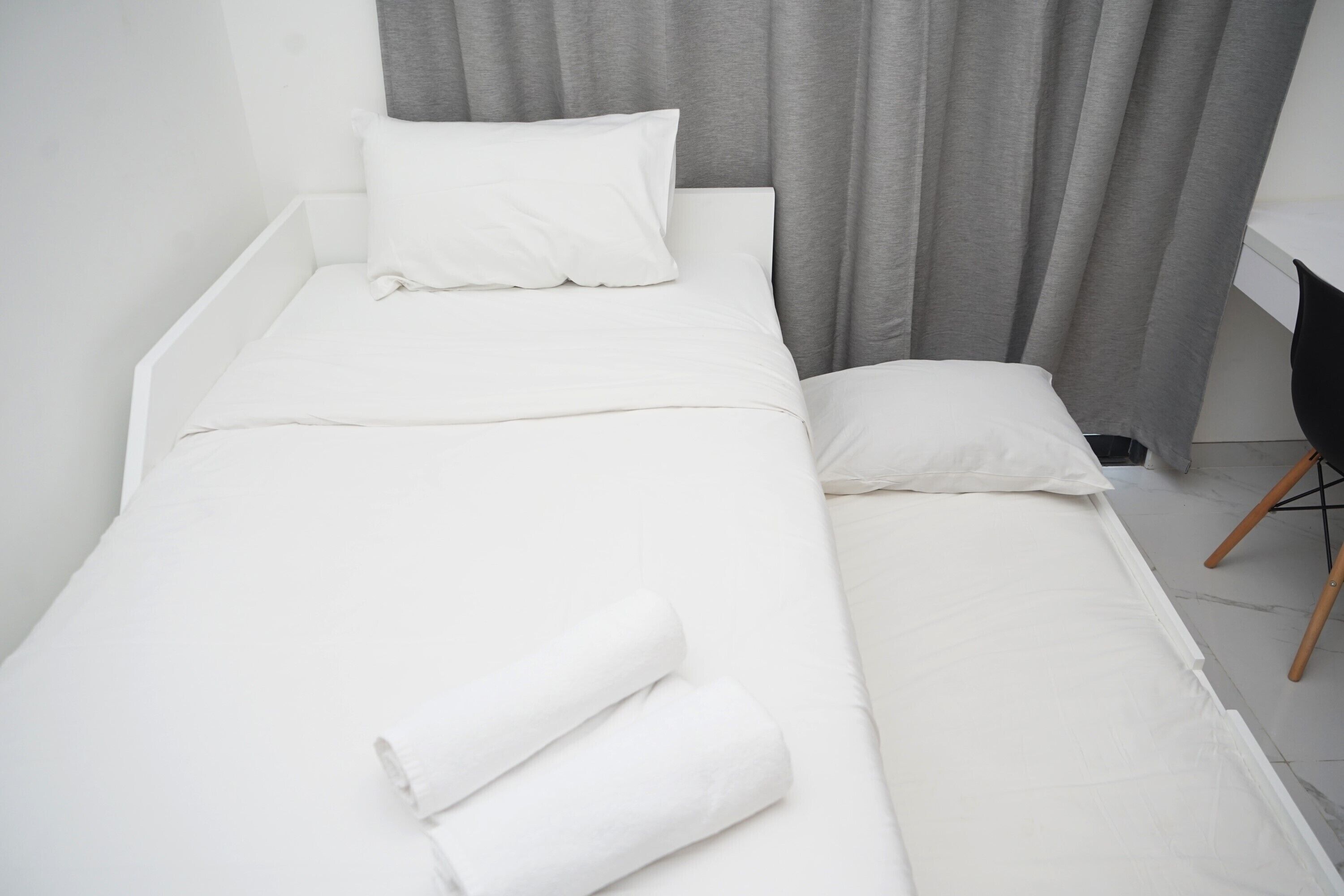 A single bed with white bedding and pillows, a grey curtain, and a black chair with wooden legs.