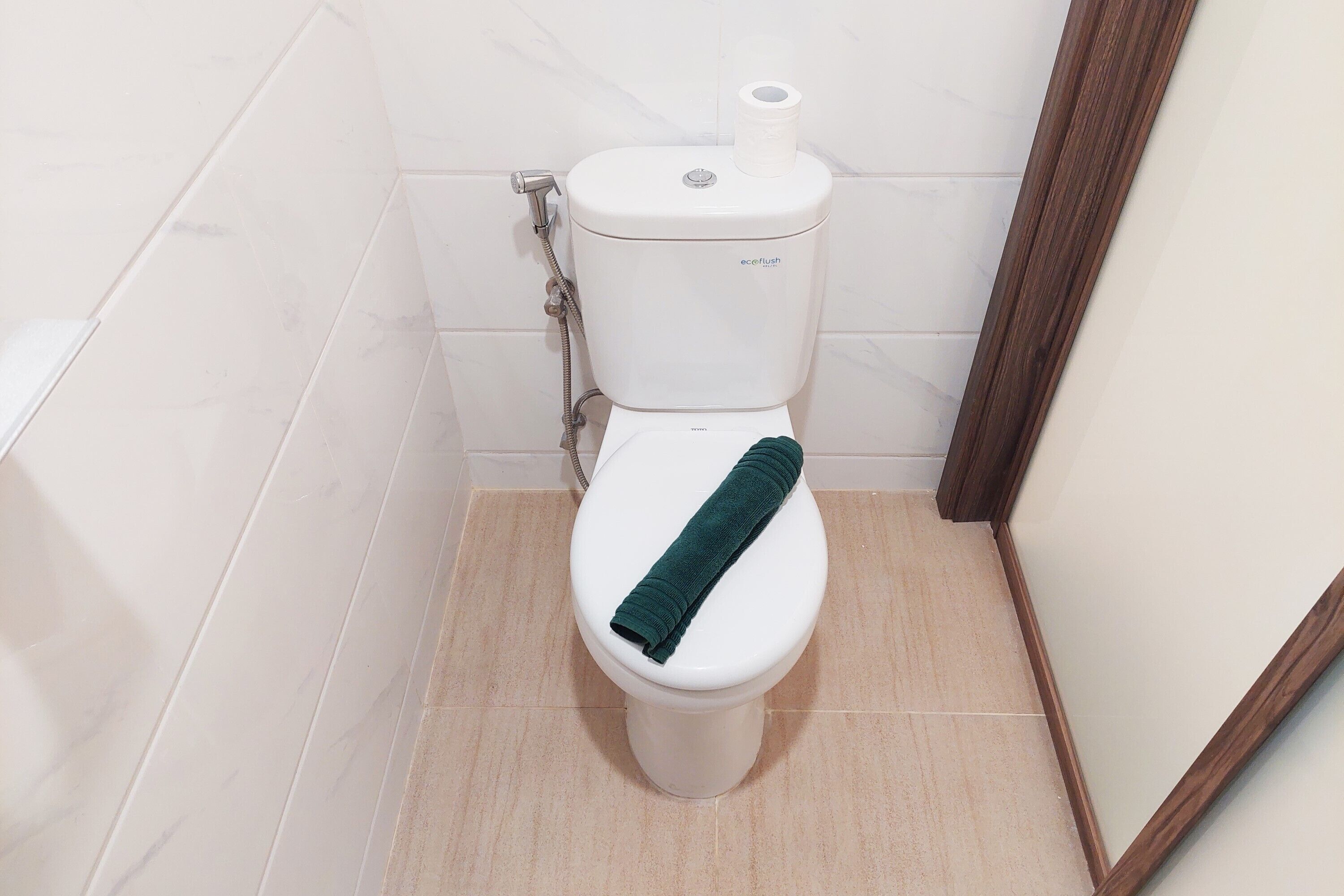 Sanitário com uma toalha verde no assento, suporte para papel higiênico e mangueira de chuveiro.