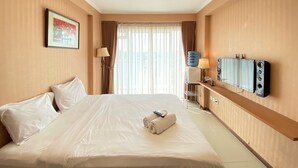A hotel room with a large bed, a flat-screen TV mounted on the wall, a desk with a lamp, and a window with curtains. - Spacious Chic Studio Room Apartment At Gateway Pasteur (Cimahi)