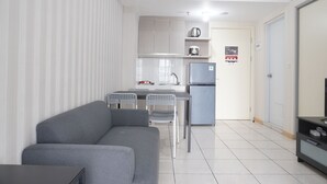 A modern living room with a grey sofa and a small dining table with two chairs. A kitchen area is visible in the background with a refrigerator, a kettle, and a coffee maker.