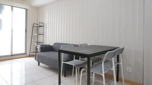 A modern living room with a grey sofa, a black table, and white chairs. There is a window with blinds, a ladder, and an air conditioning unit on the wall.