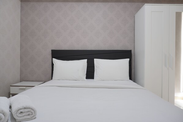 A neatly made bed with white linens and pillows, a small bedside table with a drawer, and a white wardrobe with a mirror.