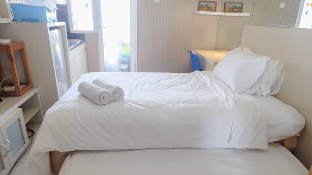Una cama bien hecha con sábanas blancas y toallas dobladas, una mesita de noche con una lámpara, y una ventana con persianas.