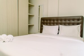 Cama bem arrumada com lençóis e almofadas brancas, cabeceira estofada marrom e um conjunto de toalhas enroladas ao lado.