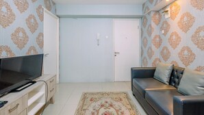 A living room with a TV on a wooden cabinet, a sofa with cushions, a patterned rug, and a door with a light switch. - Comfort And Strategic 2Br At Bassura City Apartment (Jakarta)