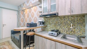 A compact kitchen with a gas stove, a coffee maker, and a sink. There is a small bar stool and a chair. - Comfort And Strategic 2Br At Bassura City Apartment (Jakarta)