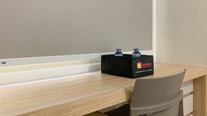 A wooden desk with a black box labeled 'Travelio' containing two water bottles, a grey chair, and a white wall. - Comfort Studio Room At Margonda Residence 5 Apartment (Depok)