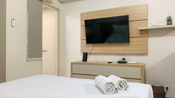 A hotel room with a bed, a TV mounted on a wooden wall, a bedside table with a speaker, and a small shelf with a plant. - Comfort Studio Room At Margonda Residence 5 Apartment (Depok)