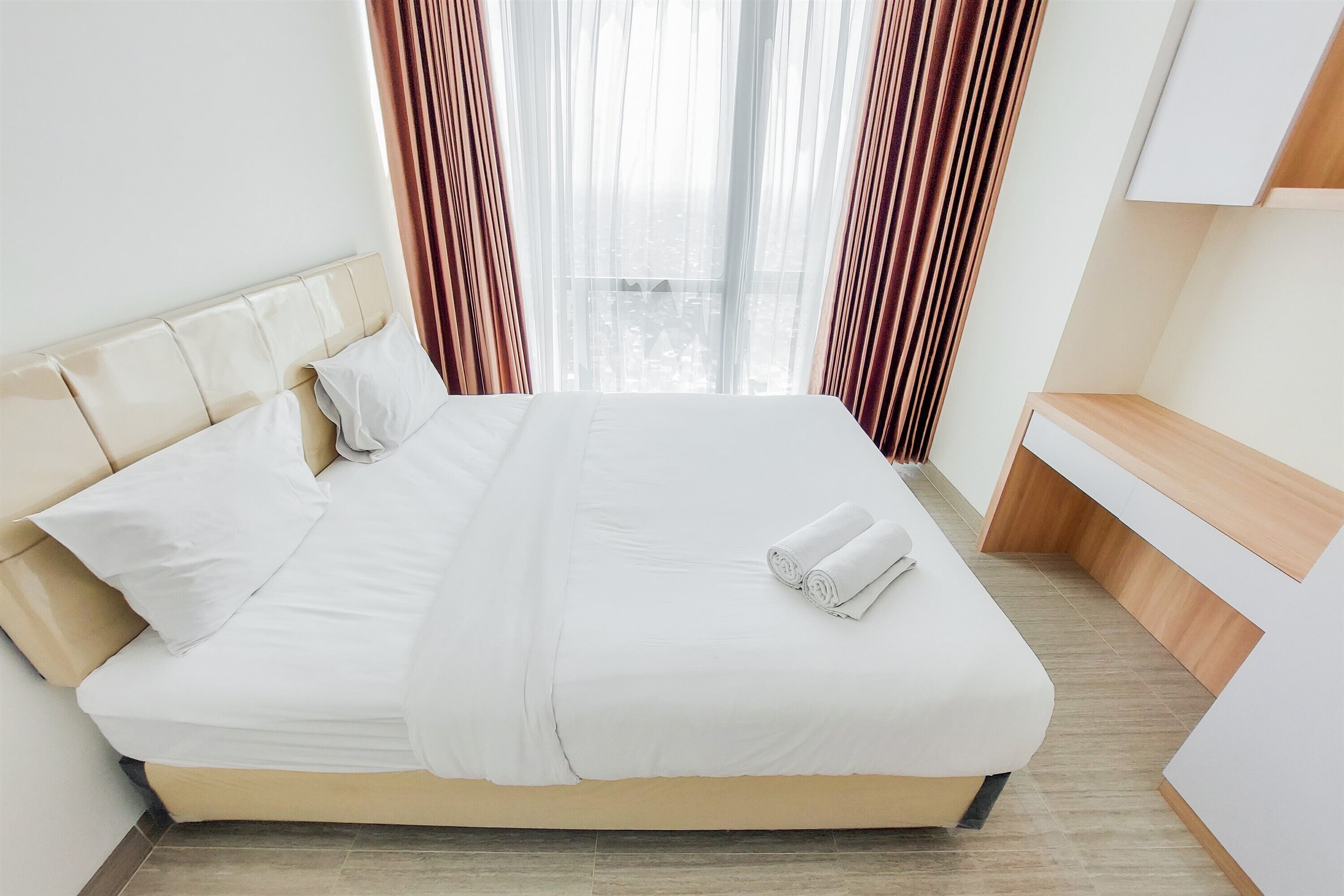 A neatly made bed with white linens and a rolled towel, a wooden nightstand, and a view of the city through sheer curtains.