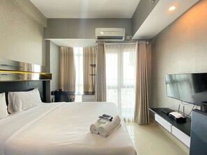 A hotel room with a large bed, a flat-screen TV, an air conditioning unit, and a desk with a computer and a router. - Minimalist And Affordable Studio Apartment At Taman Melati Jatinangor (Hegarmanah)