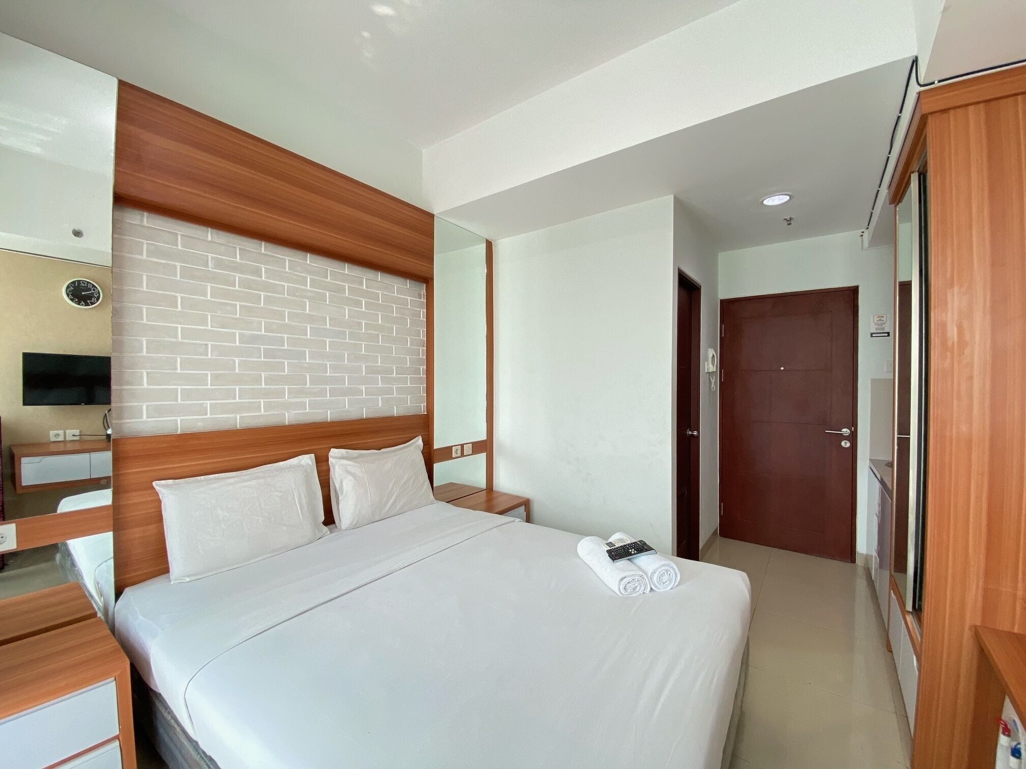 Habitación de hotel con cama, cabecera de madera, televisor de pantalla plana, mesita de noche pequeña y una puerta.