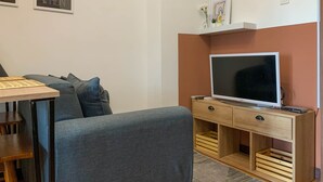 A modern living room with a grey sofa, wooden coffee table, and a TV on a wooden stand. There are wall-mounted shelves with decorative items and a framed picture. - Comfort And Nice 2Br At Cinere Resort Apartment (Depok)
