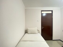 Una habitación pequeña con una cama individual, pared blanca lisa, puerta de madera oscura y almohada blanca en la cama.