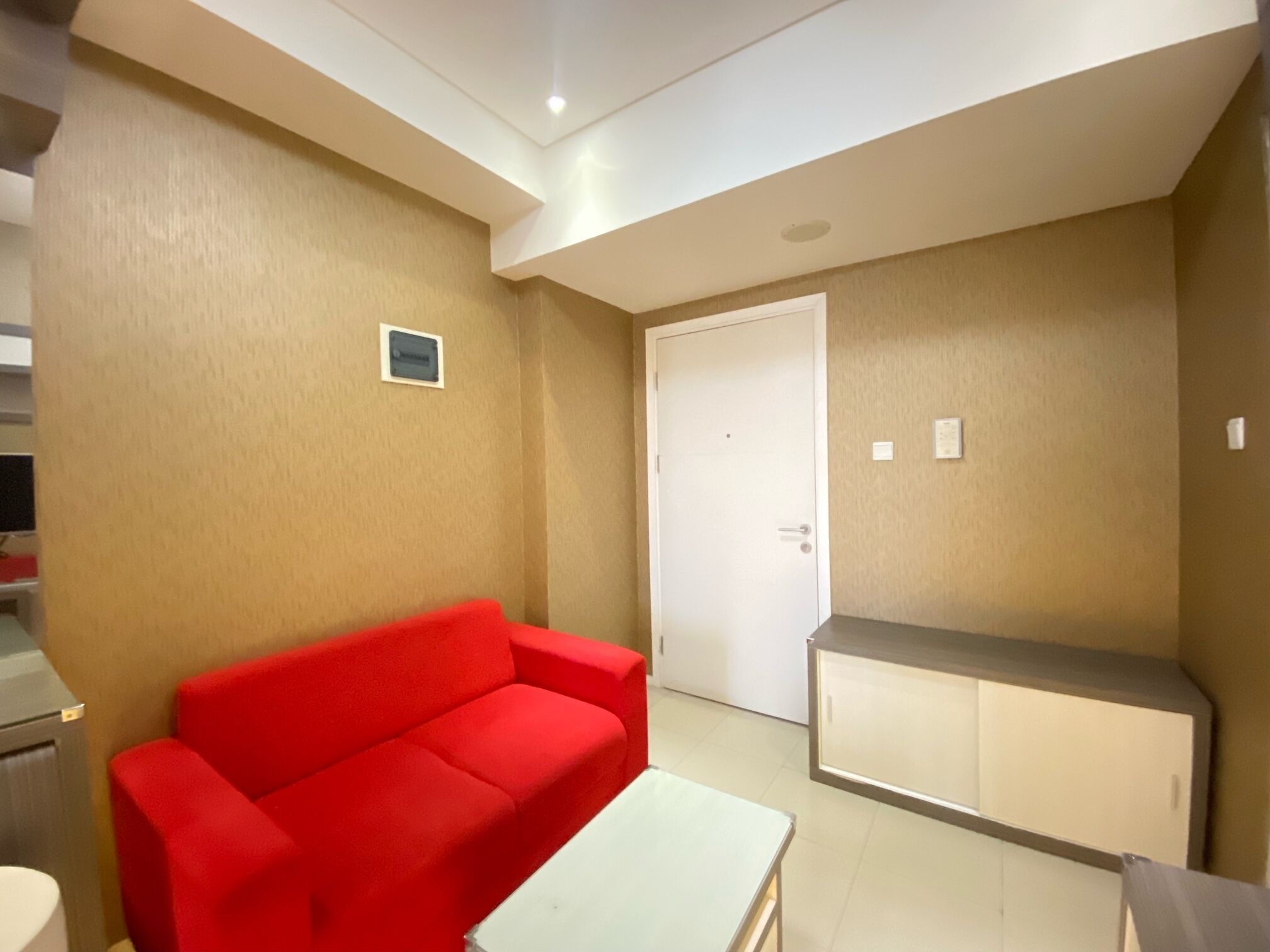 A room with a red sofa, a white table, a white door, and a kitchenette area.