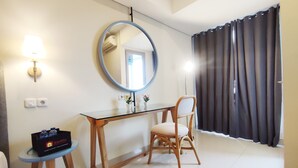 A hotel room with a wooden desk, a chair, a round mirror, a lamp, a small table with a box, and a window with curtains. - Elegant And Homey Studio At Marquis De Lafayette Apartment (Semarang)