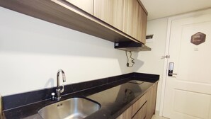 A kitchen with a black countertop, stainless steel sink, wooden cabinets, and a white door with a lock. - Elegant And Homey Studio At Marquis De Lafayette Apartment (Semarang)