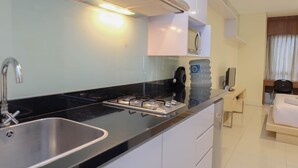 Una cocina compacta con pileta, hornalla y una pequeña mesada. Hay un dispensador de agua y un espejo sobre la mesada. Se ve una cama y un escritorio en la habitación contigua.