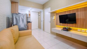 A modern living room with a sofa, a flat-screen TV mounted on a wooden wall, a small table with a power strip, and a refrigerator in the background. - Well Designed 2Br Apartment At Springlake Summarecon Bekasi (Bekasi)