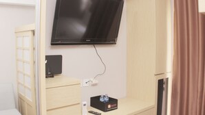 A room with a mounted flat-screen TV, a small desk with a remote control, a water bottle, and a box labeled 'travel'.